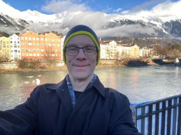 Luke Perry in a coat, scarf, and knit hat, stands in front of a body of water, buildings, and mountains while in Austria for a guest lecture.