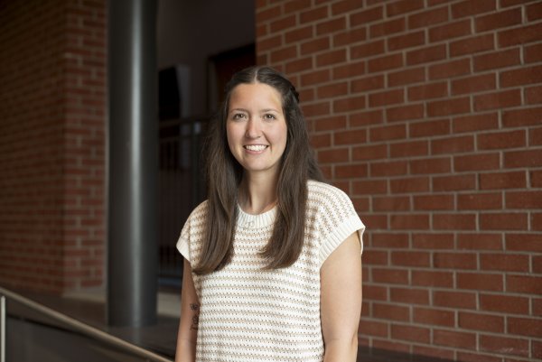 Kyla Reed, with long brown hair past her shoulders, wearing striped short sleeve shirt, stands in front of a brick wall and smiles.